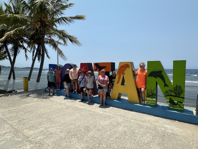 A tour group at the ROATÁN sign, palms and sea behind