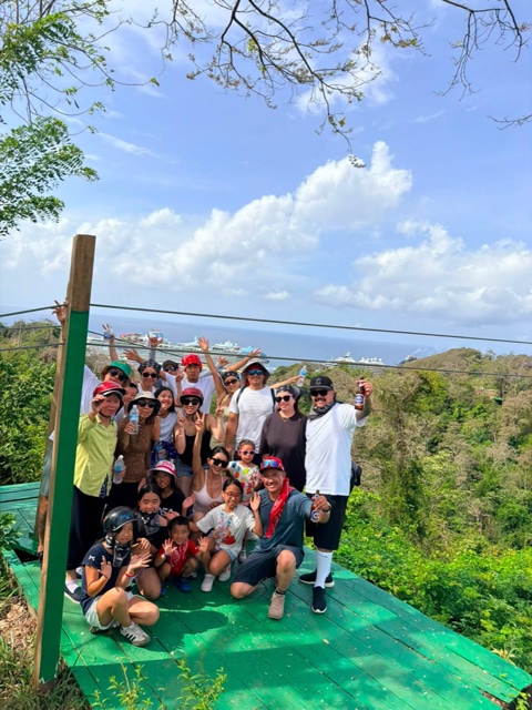 The whole zipline group on the platform, cruise ships in the bay behind